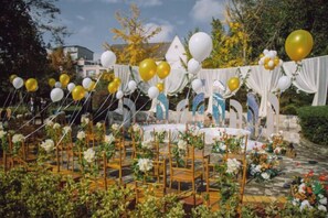 Montaje para un evento al aire libre con escenario, arreglos florales y un telón de fondo de cortinas blancas.