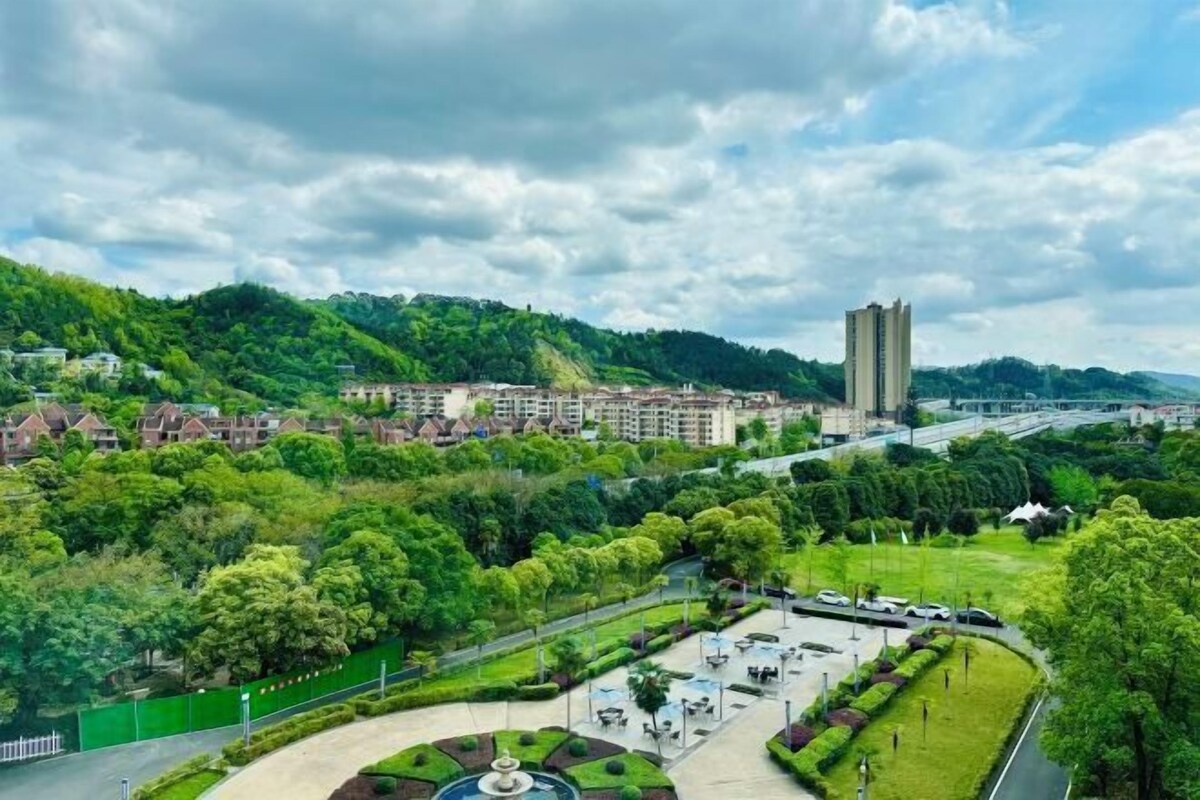 A park with a fountain, surrounded by greenery and buildings in the background.