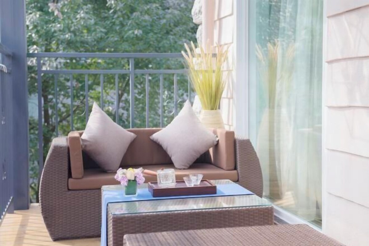 A balcony with a wicker sofa, cushions, a glass table, and a vase with flowers.