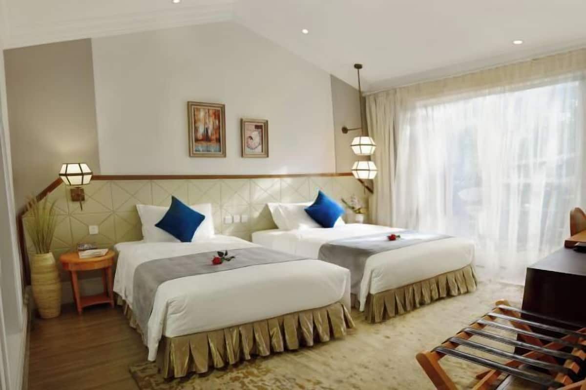 A hotel room with two beds, a small table, a chair, and a large window with sheer curtains.