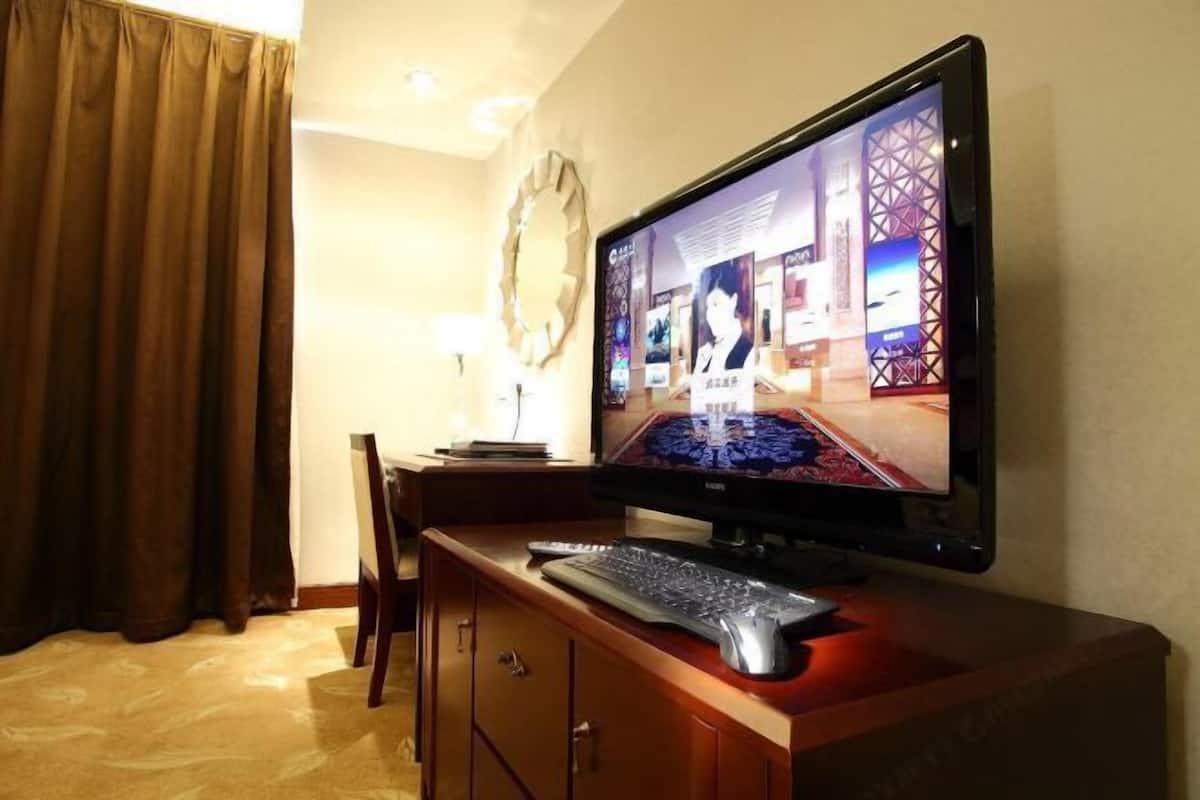 Ett hotellrum med en platt-TV på ett skrivbord i trä, en stol, en skrivbordslampa och ett stort fönster med gardiner.