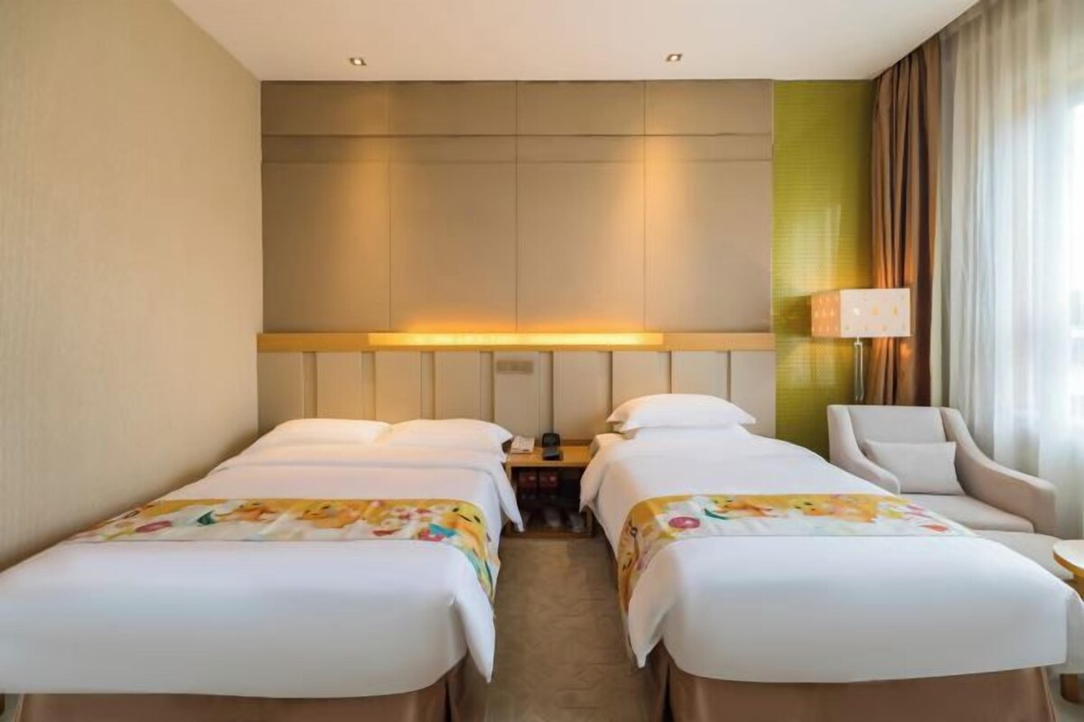 A hotel room with two beds, a desk, a chair, and a lamp.