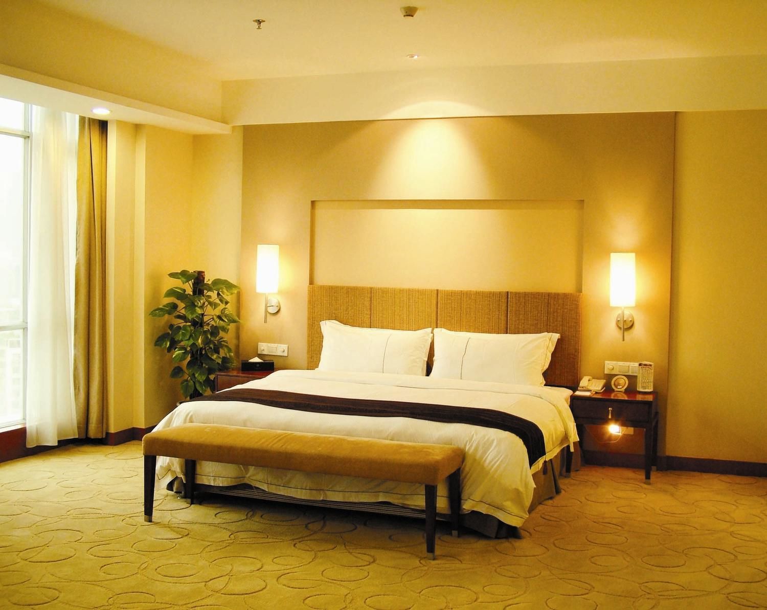 A hotel room with a large bed, a bedside table with a lamp, a nightstand with a phone, and a potted plant.