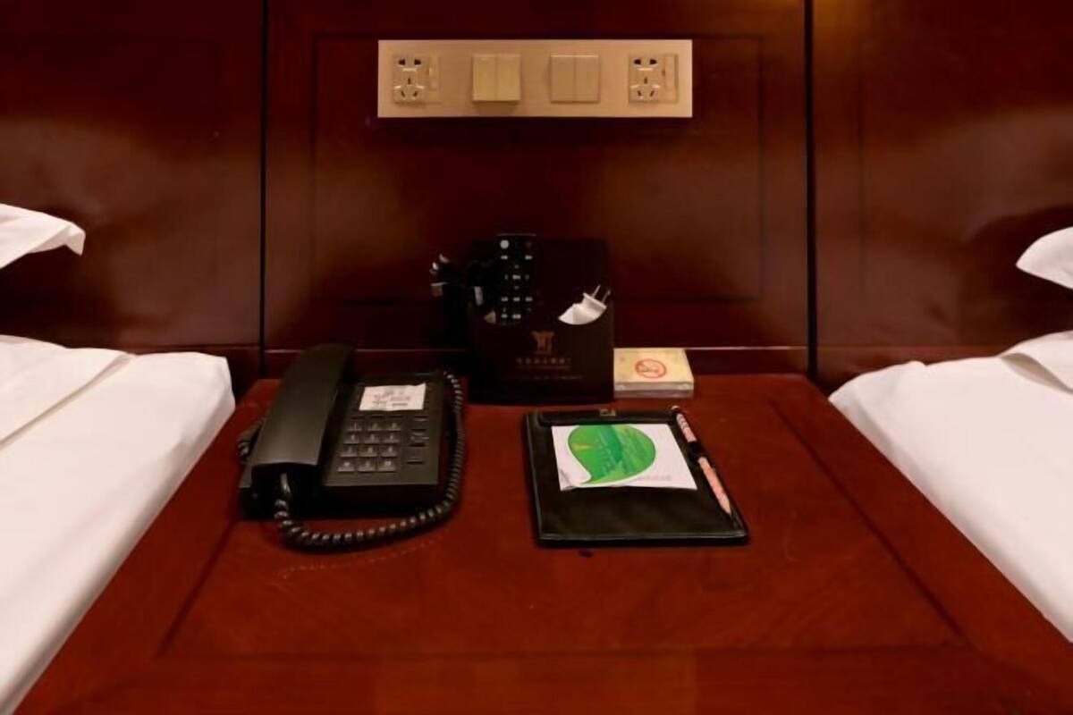 Una habitación de hotel con una cama de madera, un teléfono en la mesita de noche, un dispositivo electrónico negro, un bloc de notas y una pluma.