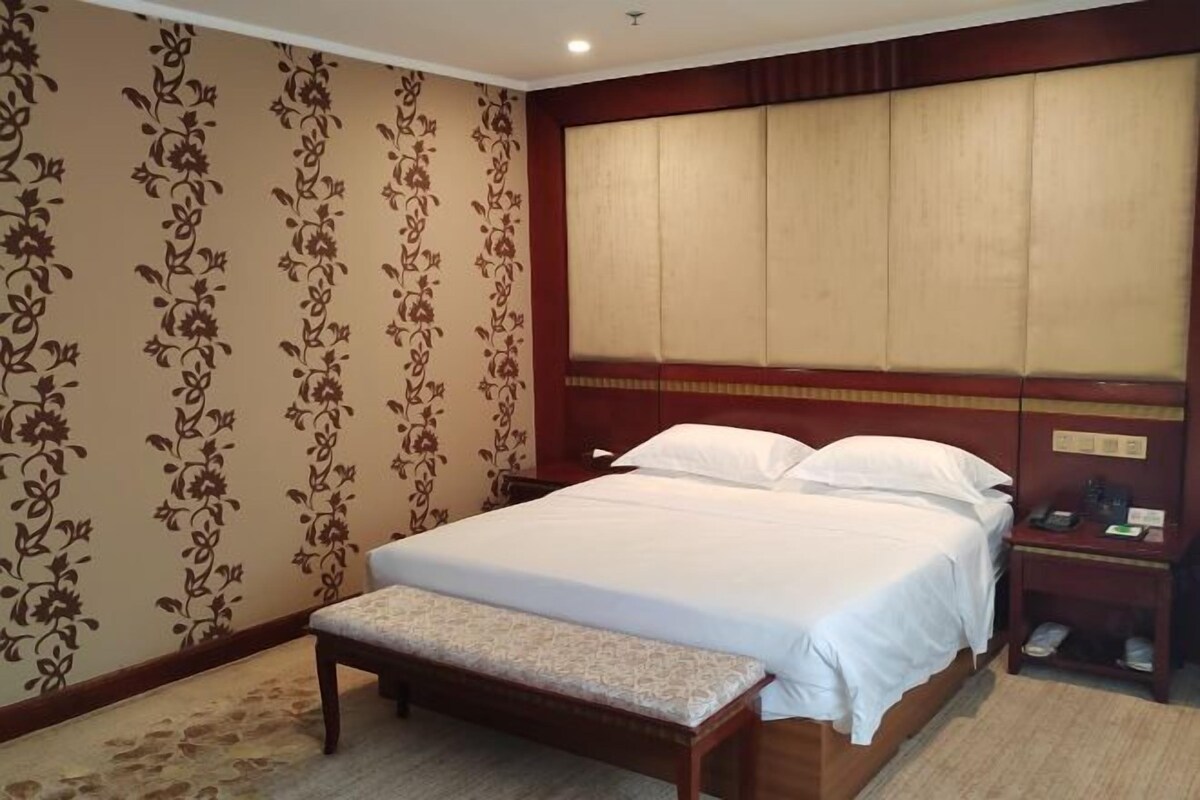 A hotel room with a bed, a nightstand, a bench, and a wall with floral wallpaper.