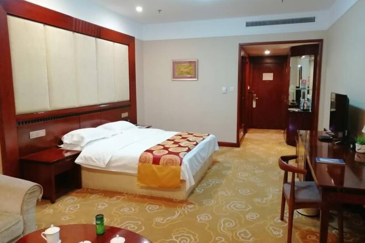 Habitación de hotel con cama, mesita de noche, una silla, una pequeña mesa con taza y platillo, y un televisor.