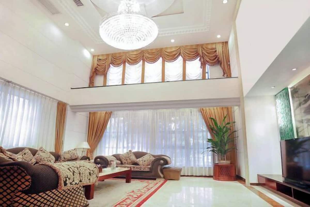 A spacious living room with a high ceiling, a chandelier, a sofa set, a television, and a large window with curtains.