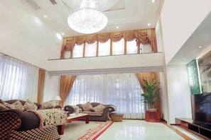 A spacious living room with a high ceiling, a chandelier, a sofa set, a television, and a large window with curtains. - Royal Prince Hotel (Quanzhou)