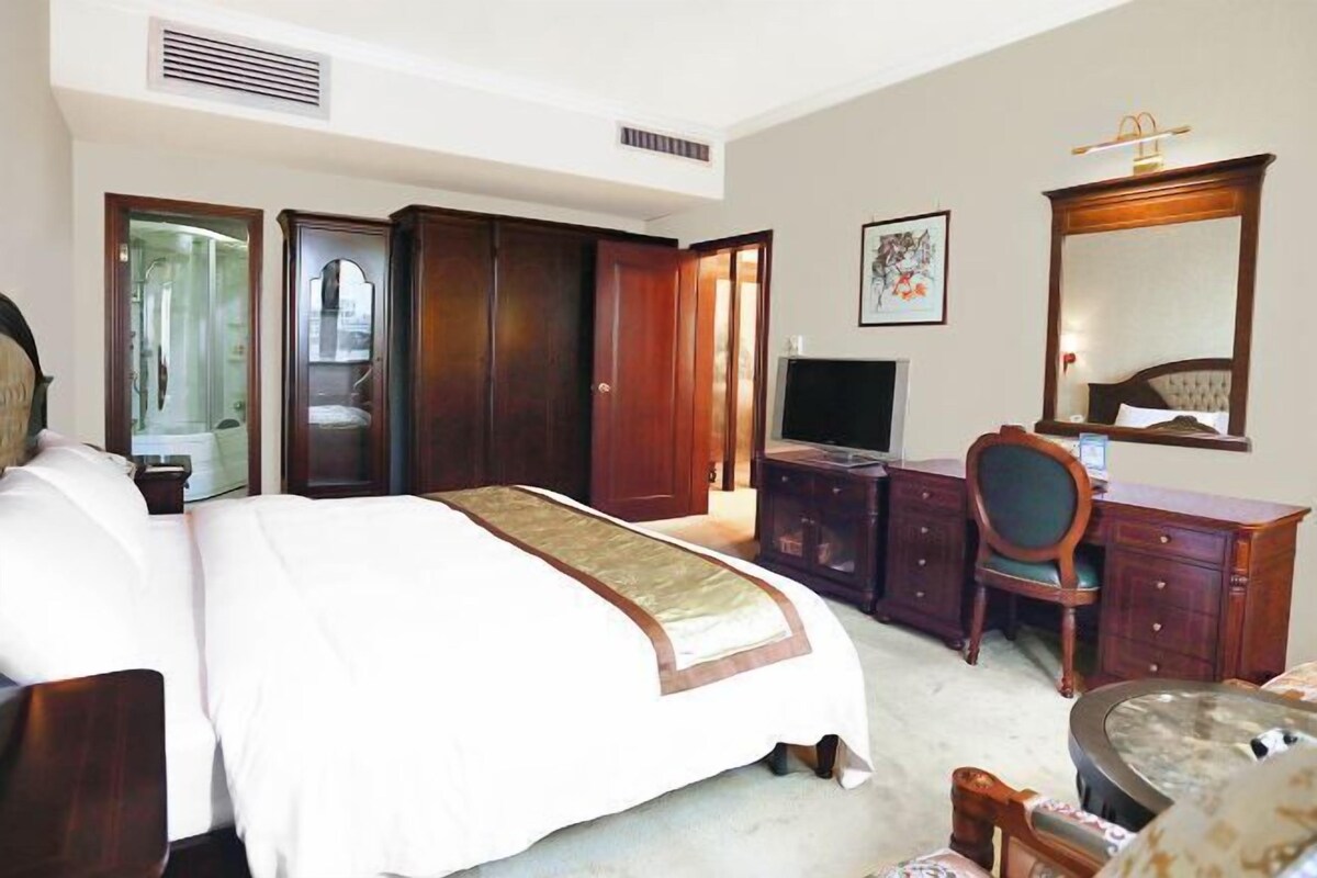A hotel room with a large bed, a desk, a chair, a television, and a wardrobe.