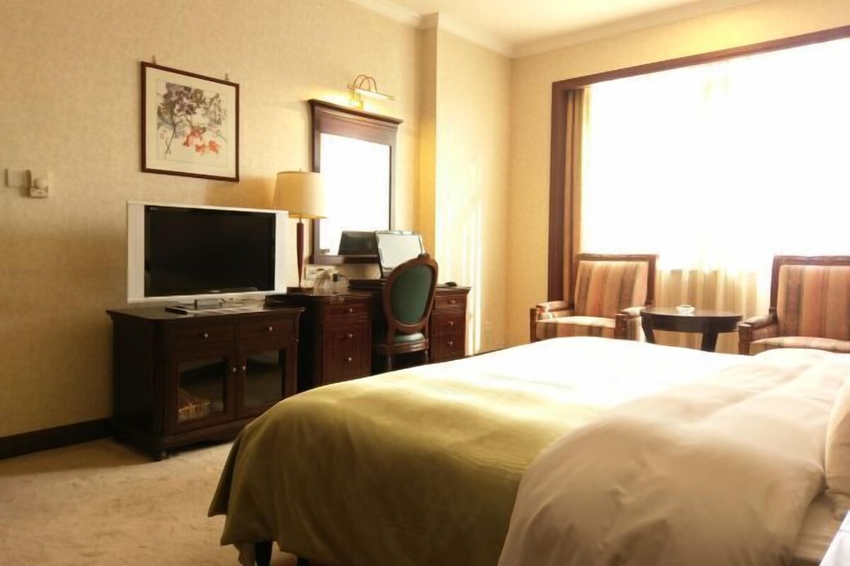 A hotel room with a bed, a desk with a television, a chair, a lamp, and a large window.