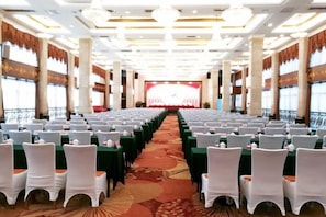 A conference room with rows of tables and chairs, a stage at the front, and large windows along the sides. - Royal Prince Hotel (Quanzhou)