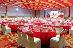 Un salón de banquetes con mesas redondas preparadas para eventos, sillas blancas con cintas rojas y un suelo cubierto de alfombra roja.