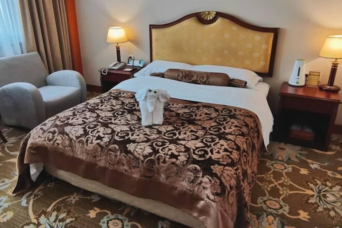 Ein Hotelzimmer mit einem großen Bett, einem Sessel, zwei Nachttischen mit Lampen und einem gemusterten Teppich.