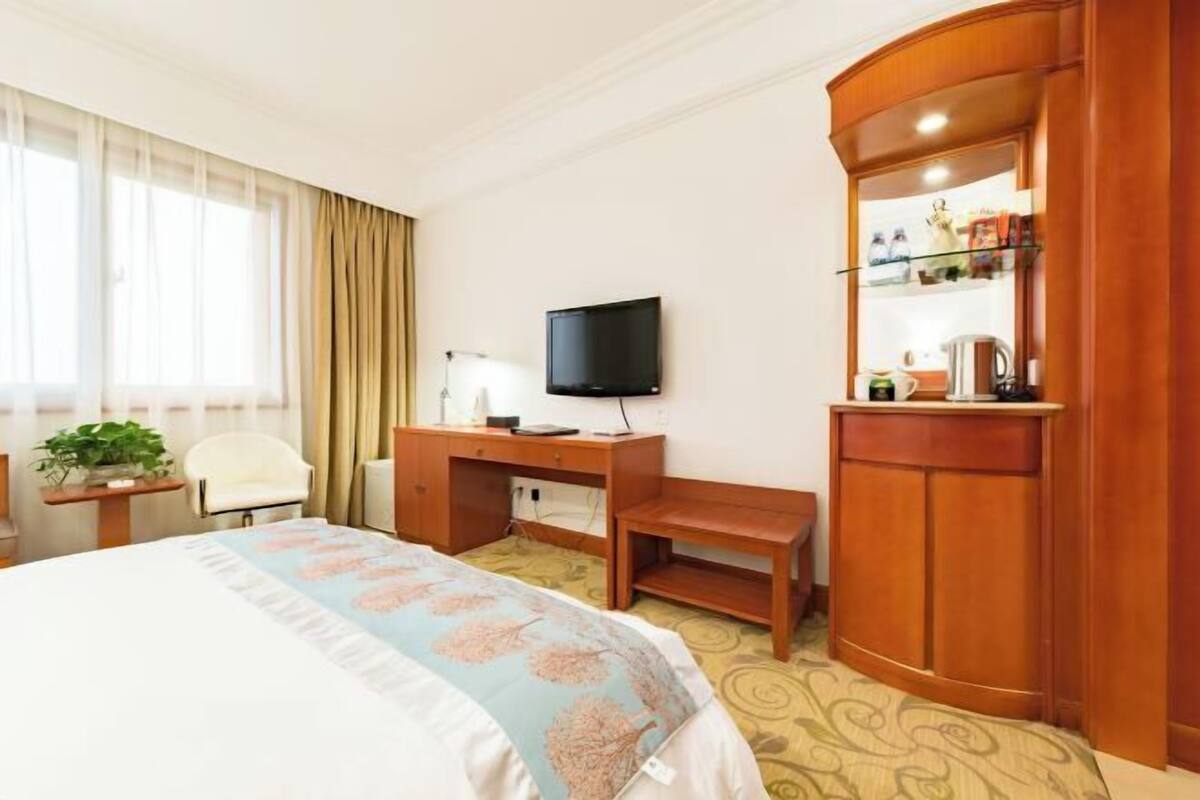 Ein Hotelzimmer mit einem Bett, einem Schreibtisch mit Fernseher, einem Sessel, einem kleinen Tisch mit Lampe und einer Minibar.