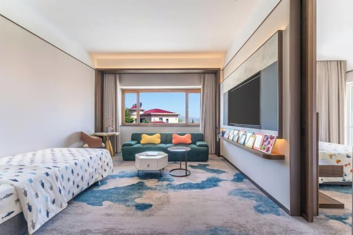Een hotelkamer met een bed, een slaapbank, een tafeltje, een televisie en een raam met uitzicht op gebouwen.