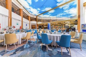 A spacious dining area with round tables and chairs, a high ceiling with a sky-like design, and a view of the adjacent restaurant. - Tarshan Hotel (WEIHAI)