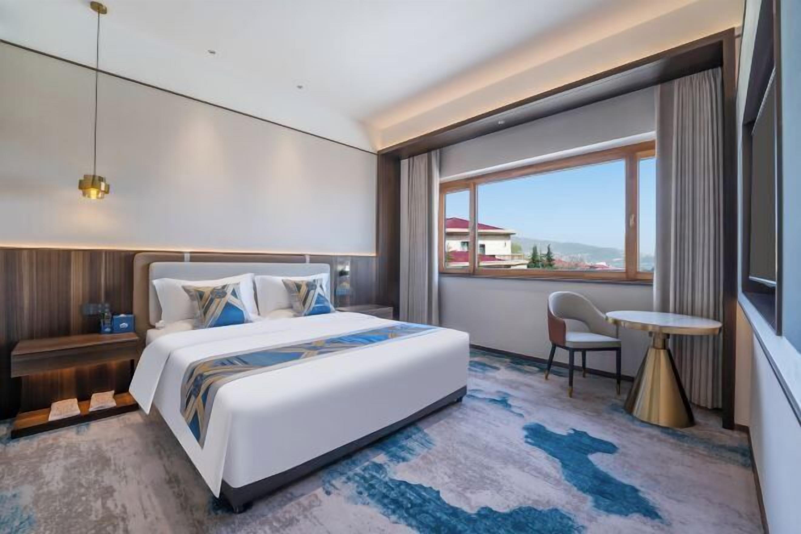 Habitación de hotel moderna con una cama grande, una mesita pequeña, una silla y un ventanal con vistas a edificios.