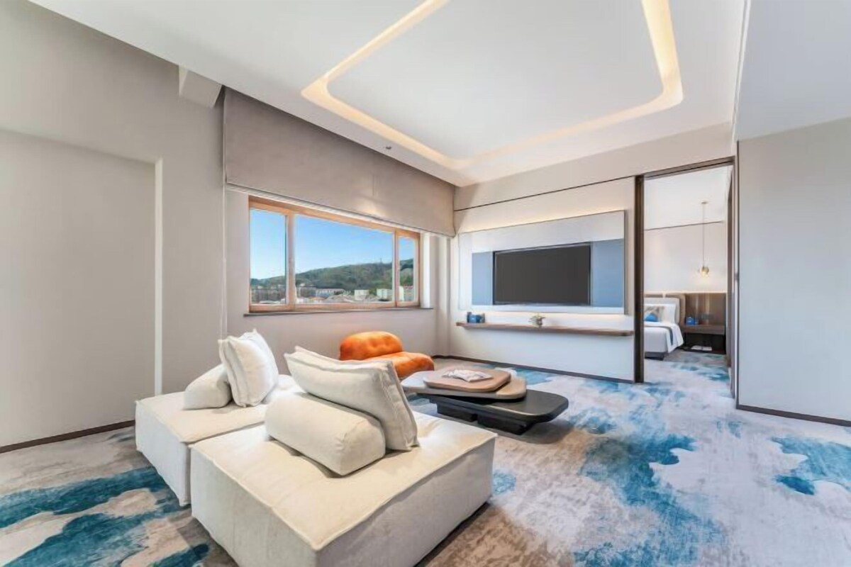 Een moderne hotelkamer met een groot bed, een zithoek met een bank en poef, een flatscreen-tv en uitzicht op het omliggende landschap.