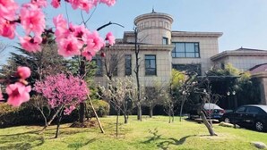 A building with a circular tower, surrounded by a well-maintained garden with various trees and shrubs. - Tarshan Hotel (WEIHAI)