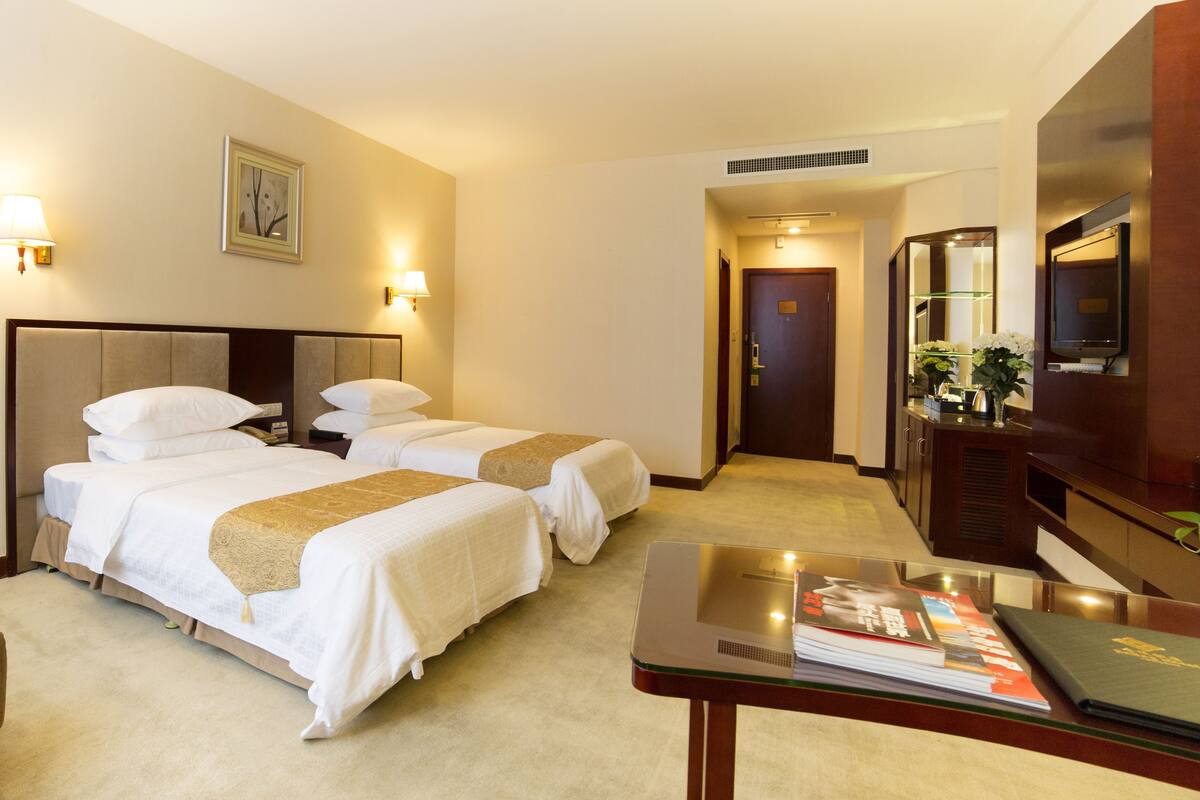 Een hotelkamer met twee bedden, een televisie, een bureau met een tijdschrift en een deur naar een gang.