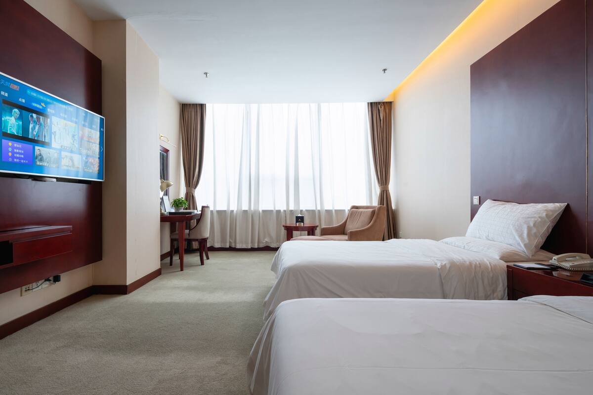 Een hotelkamer met twee bedden, een televisie aan de muur, een bureau met een stoel en een raam met gordijnen.
