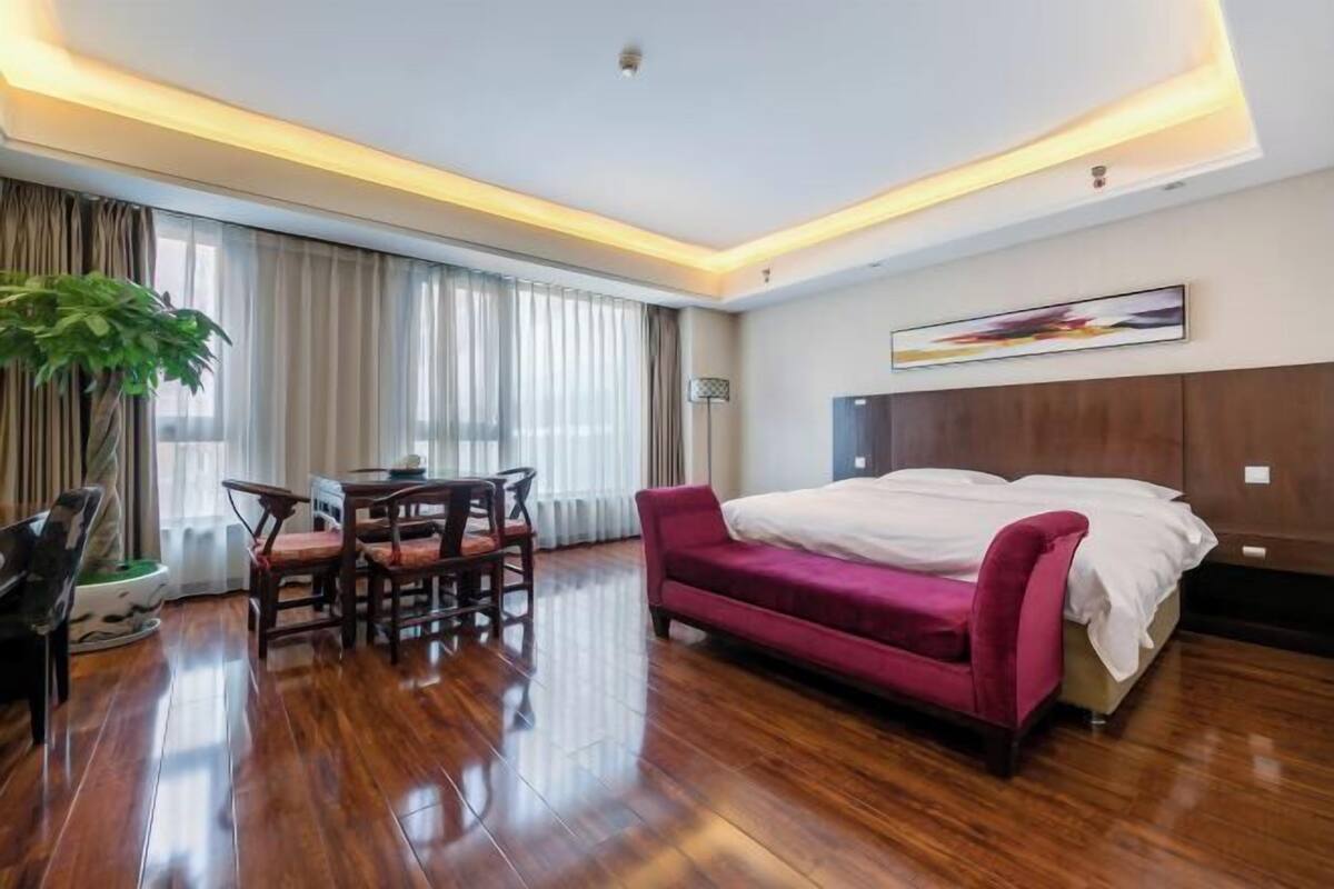 A modern hotel room with a wooden floor, a large bed with a headboard, a dining table with chairs, and a potted plant.