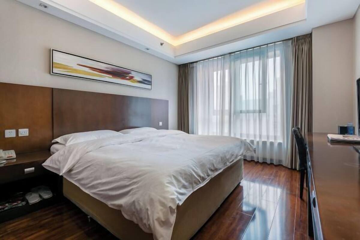 A hotel room with a bed, a desk, a chair, a window with curtains, and a painting on the wall.