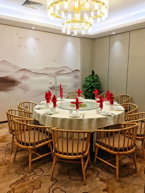 Un coin repas avec une table ronde dressée pour six personnes, entourée de chaises en osier, un lustre au plafond et une fresque murale.