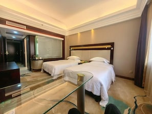 A hotel room with two beds, a glass table, a TV, and a large window.