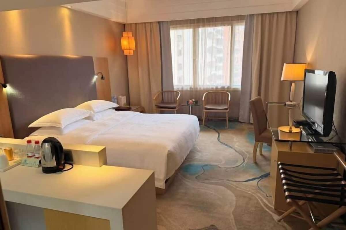 Ein Hotelzimmer mit einem Bett, einem Schreibtisch mit Fernseher, einem Sessel, einem kleinen Tisch und einem Fenster mit Vorhängen.