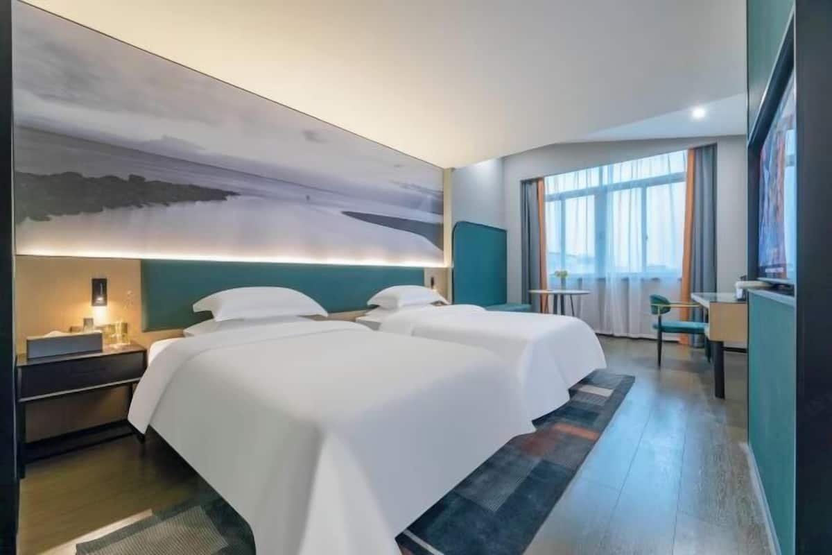 A hotel room with two beds, a large painting on the wall, a desk with a chair, and a window with curtains.