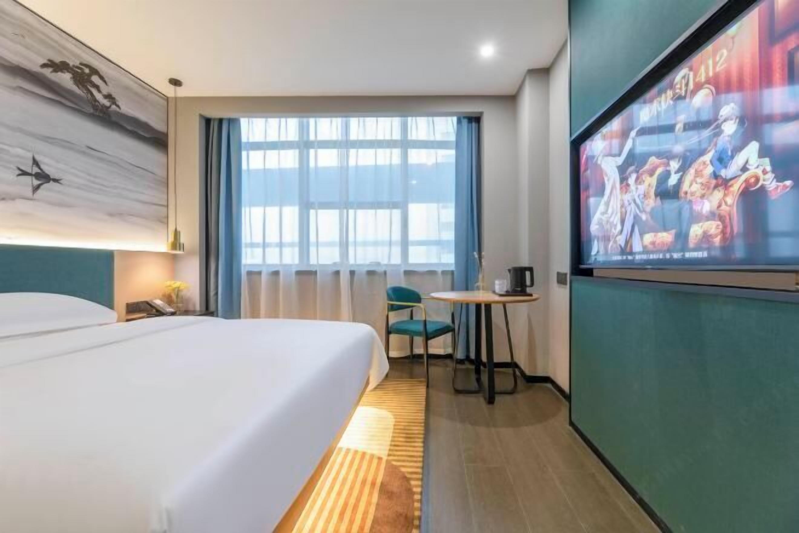 A hotel room with a bed, a desk, a chair, a TV screen, and a large window with curtains.