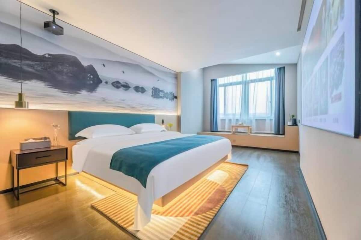 A modern hotel room with a large bed, a nightstand, a desk, a large window, and a wall-mounted projector.
