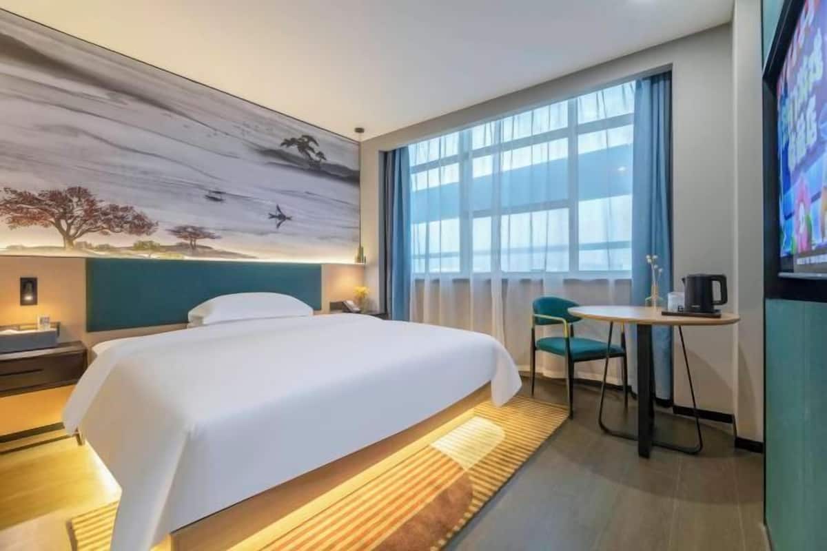 A hotel room with a bed, a desk, a chair, a large window, and a painting on the wall.