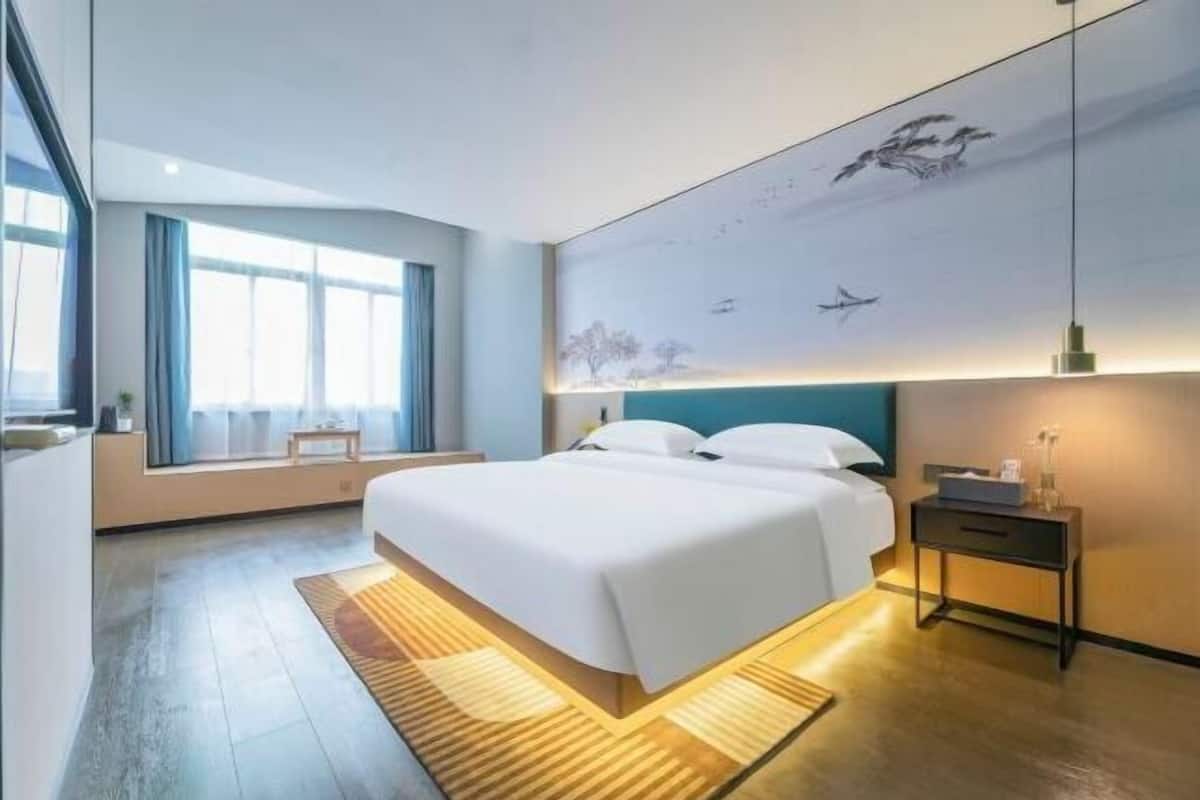 A modern hotel room with a large bed, a bedside table, a nightstand, a window with curtains, and a wall mural.