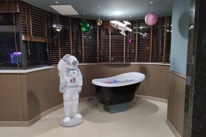 Une salle de bain avec une baignoire, une figurine d’astronaute sur le thème de l’espace et une fenêtre avec des stores.