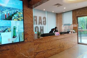 Zona de recepción de hotel con un mostrador de mármol, una pantalla digital grande que muestra paisajes urbanos y vistas al exterior a través de la ventana.