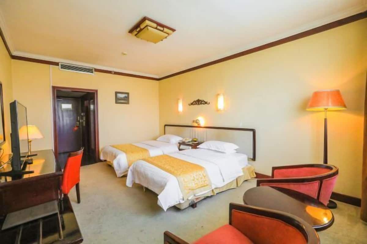 A hotel room with two beds, a desk with a chair, a lamp, and a red chair.