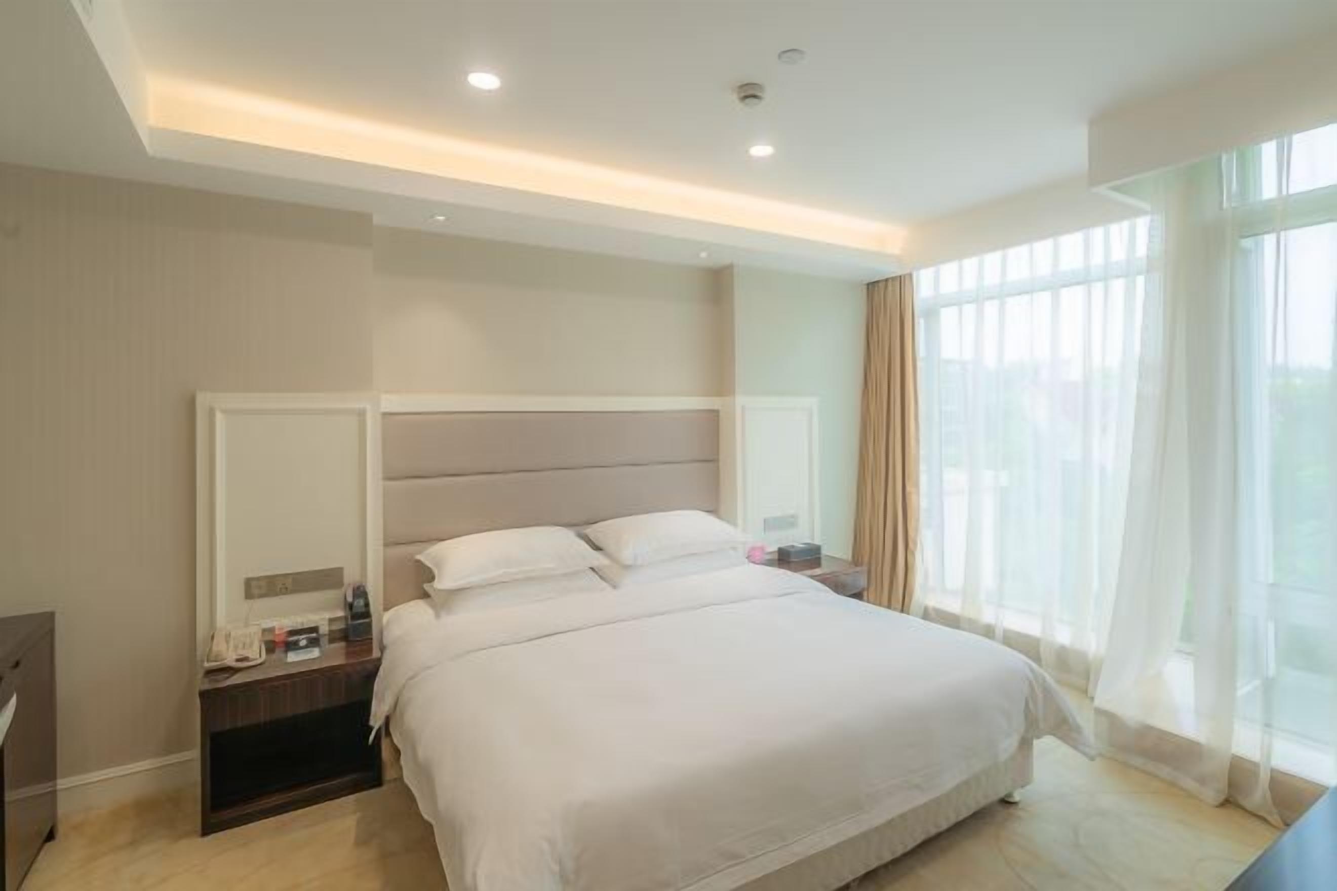 A modern hotel room with a large bed, a nightstand, a mirror, and a window with sheer curtains.