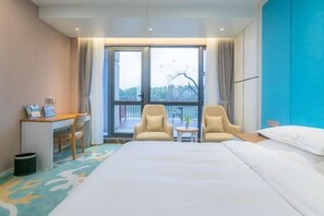 A hotel room with a large bed, two armchairs, a small table, and a desk with a computer and papers.