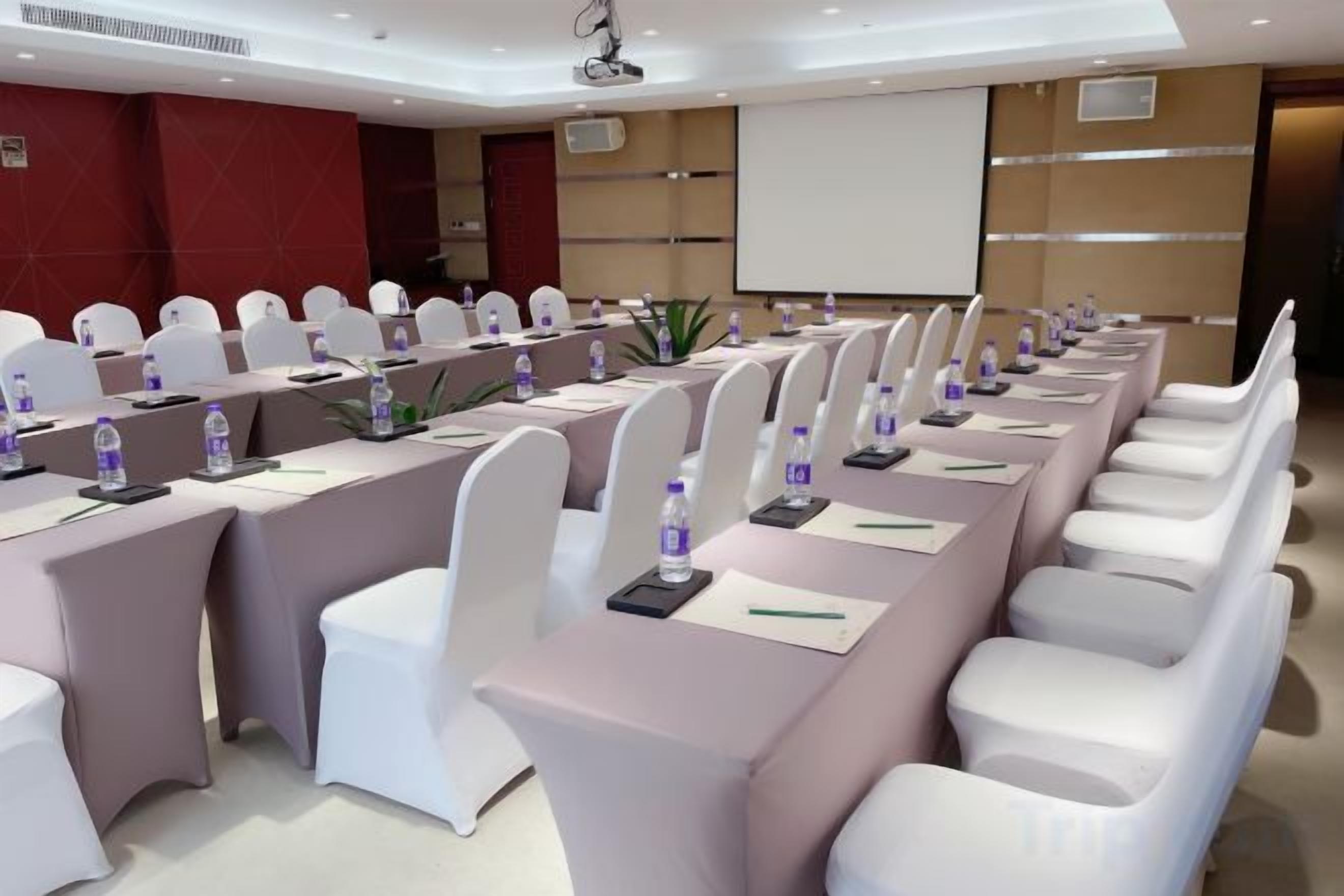 A conference room with rows of white chairs, tables with water bottles, and a projector screen.