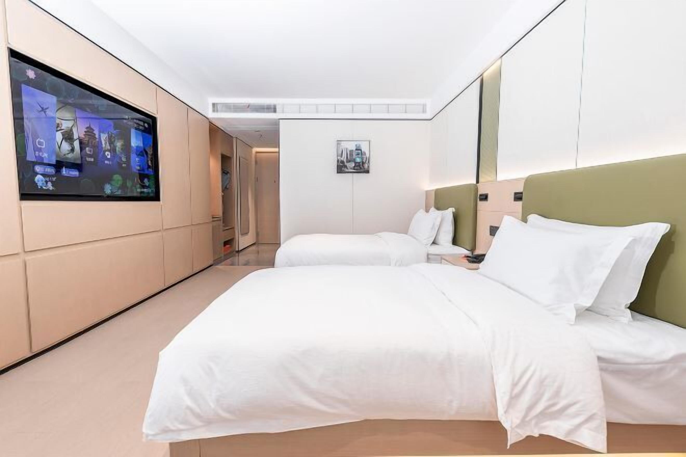 A hotel room with a large flat-screen TV mounted on the wall, two beds with white bedding, a small bedside table, and a framed picture on the wall.