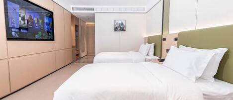A hotel room with a large flat-screen TV mounted on the wall, two beds with white bedding, a small bedside table, and a framed picture on the wall.