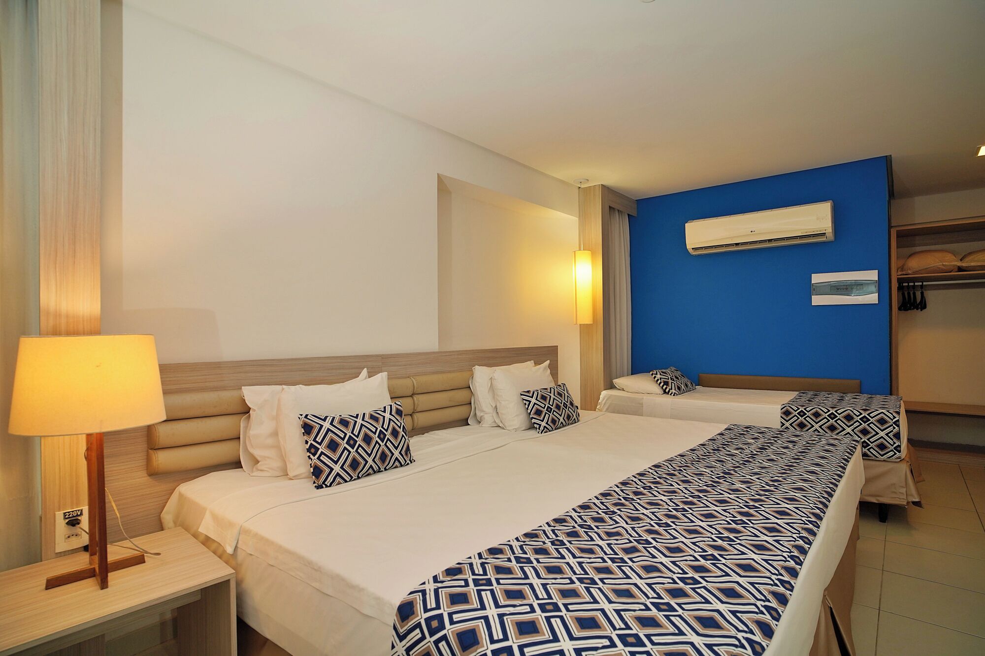 A hotel room with two beds, a bedside lamp, an air conditioning unit, and a blue accent wall.