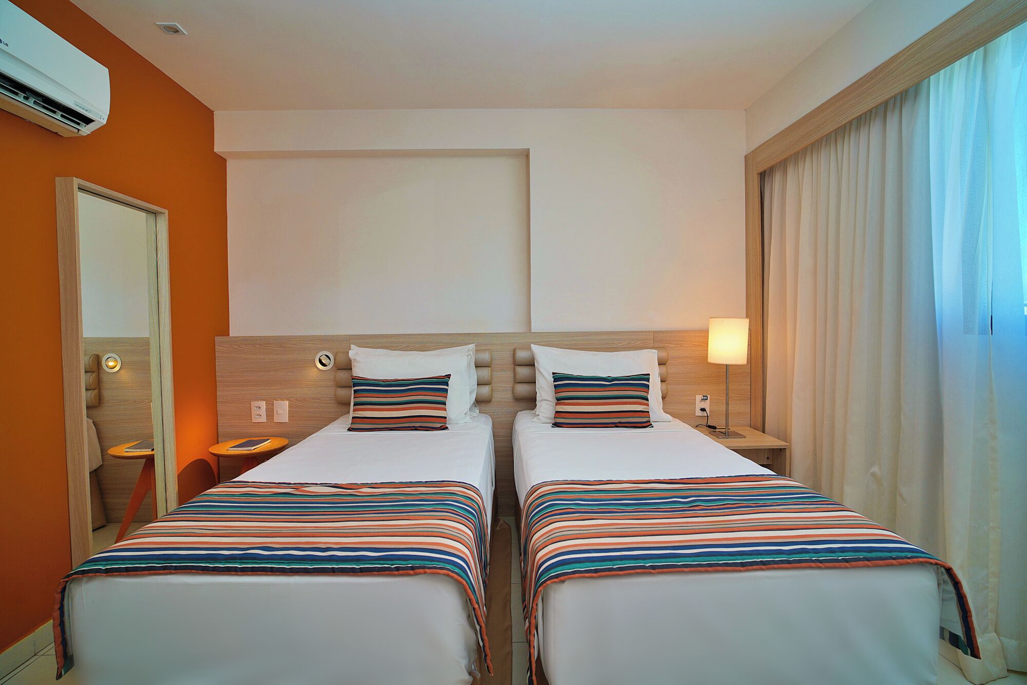 A hotel room with two beds, a wooden headboard, a nightstand with a lamp, an air conditioning unit, and a large window with curtains.