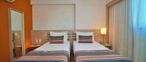 A hotel room with two beds, a wooden headboard, a nightstand with a lamp, an air conditioning unit, and a large window with curtains.