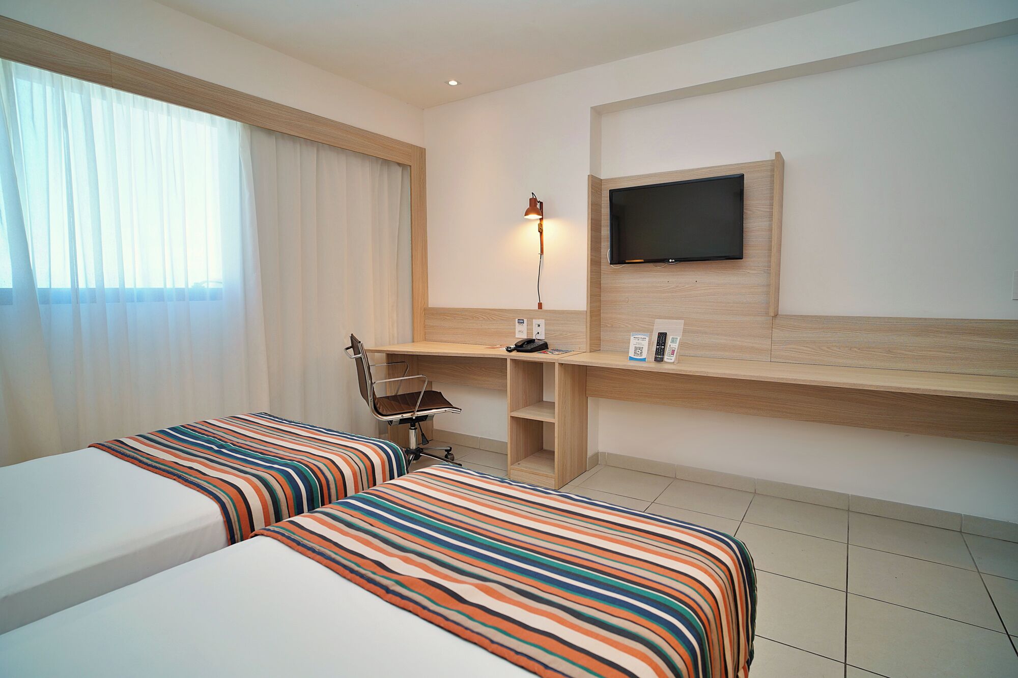 A hotel room with two beds, a desk, a chair, a TV, and a lamp.