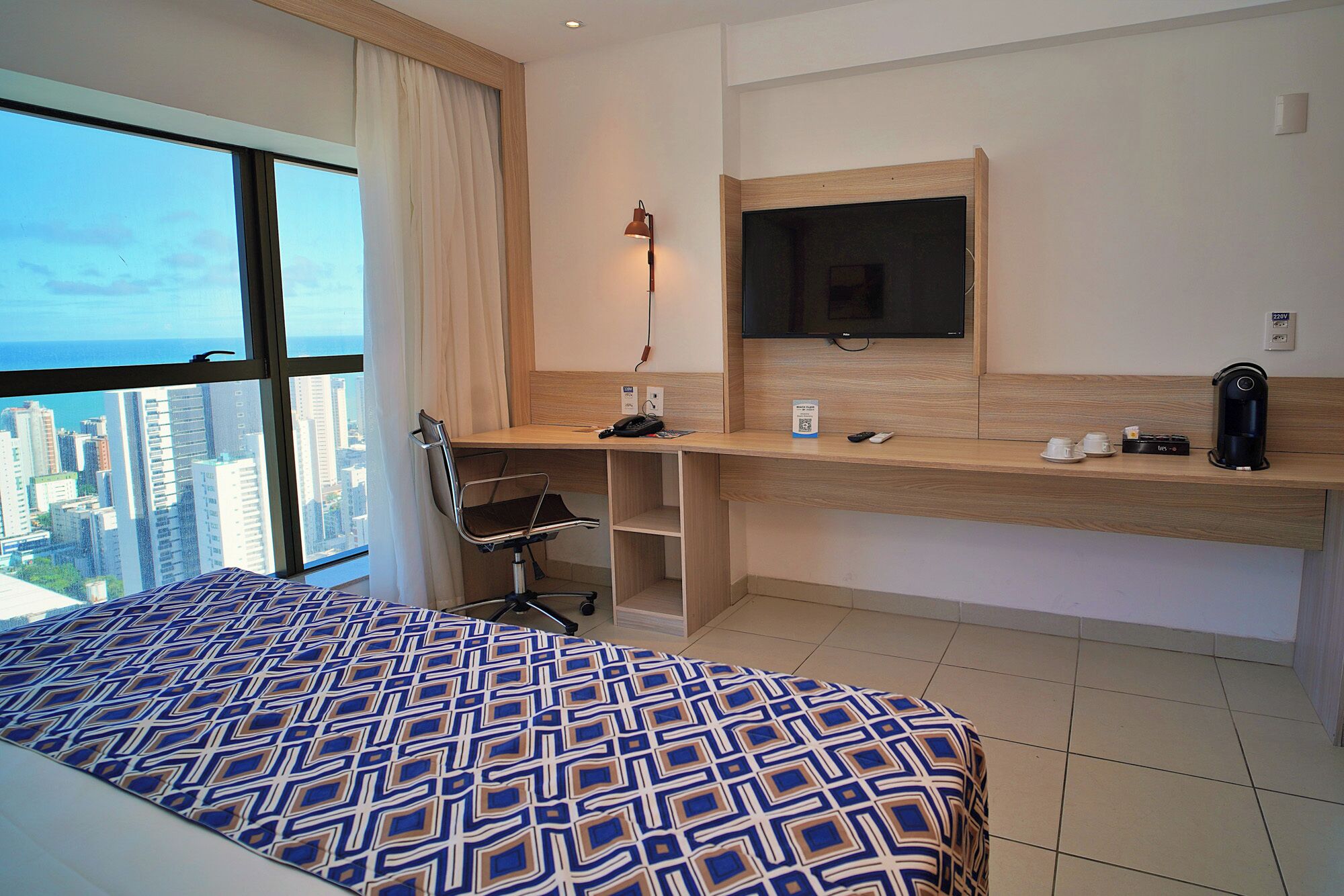 A hotel room with a bed, a desk with a computer, a TV, and a view of the city.