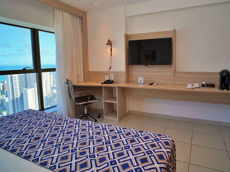A hotel room with a bed, a desk with a computer, a TV, and a view of the city.