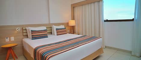Habitación de hotel con una cama grande, una mesita de noche, una lámpara y vistas al cielo a través de la ventana.